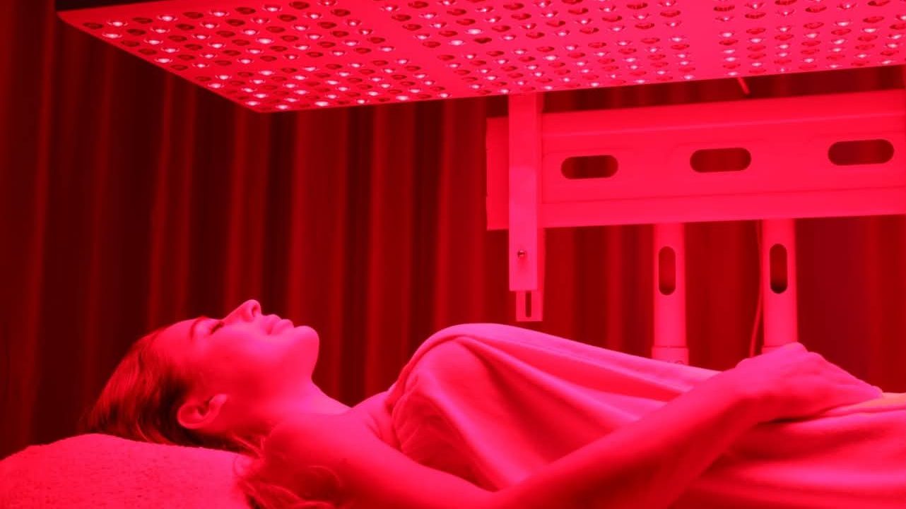 A client resting under the horizontally mounted red light panel, covered with a blanket on the treatment table
